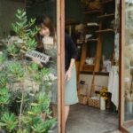 Positive young Asian female florist in apron standing near entrance and turning signboard on glass wall while working in modern floristry shop